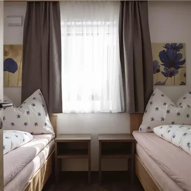 Ein einfacher, heller Raum mit zwei Einzelbetten und einer großen Fensterfront. Die Wände sind mit farbigen Bildern dekoriert, und Vorhänge sorgen für eine gemütliche Atmosphäre.