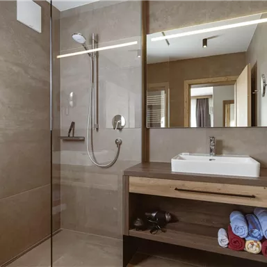 Ein modernes Badezimmer mit einer großzügigen Dusche und einem eleganten Waschbecken. Farbenfrohe Handtücher sind ordentlich auf einem Holzregal aufgereiht.