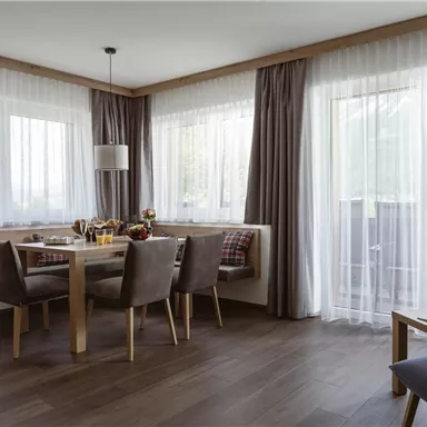 Ein modernes Esszimmer mit einem Holztisch und gemütlichen Stühlen. Große Fenster lassen viel Tageslicht herein und bieten einen Blick nach draußen.