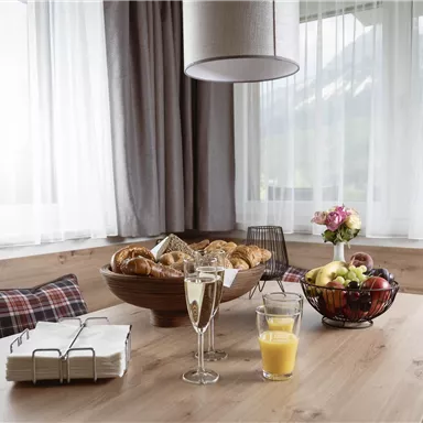 Ein einladender Frühstückstisch mit frischem Gebäck, Obst und Gläsern. Der Raum hat große Fenster mit Vorhängen und bietet eine gemütliche Atmosphäre.