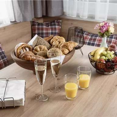 Ein gedeckter Tisch mit einer Auswahl an frischem Gebäck, Früchten und Getränken. Im Hintergrund sind gemütliche Kissen zu sehen.