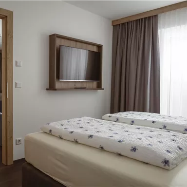 Ein gemütliches Schlafzimmer mit zwei Betten und einem Fernseher an der Wand. Die Fenster sind mit Vorhängen bedeckt und lassen viel Licht herein.