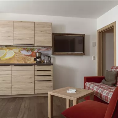 Eine moderne Küche mit Holzmöbeln und einem Fernseher an der Wand. Im Vordergrund steht ein rotes Sofa und ein kleiner Couchtisch.