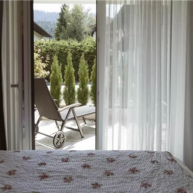 Ein gemütliches Schlafzimmer mit einem Blick auf eine Terrasse. Im Hintergrund sind grüne Pflanzen und ein Liegestuhl zu sehen.