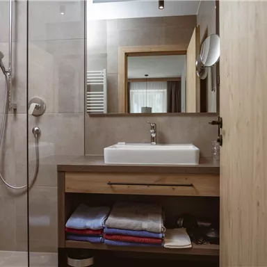 Ein modernes Badezimmer mit einer Dusche und einem Waschbecken. Handtücher sind ordentlich auf einem Holzregal gestapelt.