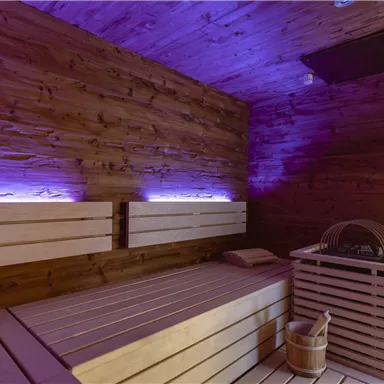 Eine moderne Sauna mit Holzverkleidung und sanfter, lila Beleuchtung. Die Bänke sind aus hellem Holz und schaffen eine entspannende Atmosphäre.