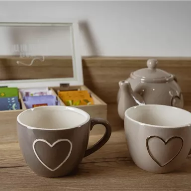 Zwei dekorative Tassen mit Herzen und eine Teekanne stehen auf einem Holzuntergrund. Im Hintergrund befindet sich eine Box mit verschiedenen Teebeuteln.