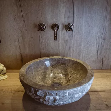 Ein elegenes Waschbecken aus Stein auf einer Holzoberfläche. Im Hintergrund sind stilvolle Wasserhähne sichtbar.