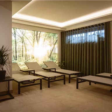 Ein entspannender Raum mit Liegen und großem Fenster, das einen Wald mit Sonnenlicht zeigt. Die Atmosphäre ist ruhig und einladend.