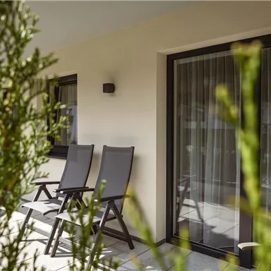 Eine gemütliche Terrasse mit zwei Liegen und großen Fenstern. Die Umgebung ist mit Pflanzen geschmückt.
