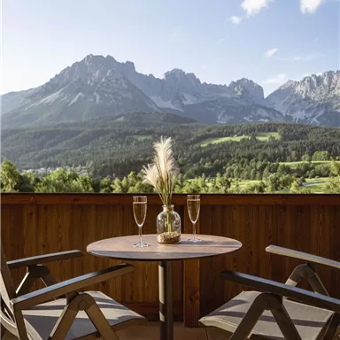 Ein gemütlicher Balkon mit einem runden Tisch und zwei Stühlen. Im Hintergrund sind majestätische Berge und ein klarer Himmel zu sehen.