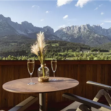 Ein Tisch auf einem Balkon mit zwei Gläsern und einer dekorativen Vase. Im Hintergrund sind beeindruckende Berge und eine malerische Landschaft zu sehen.