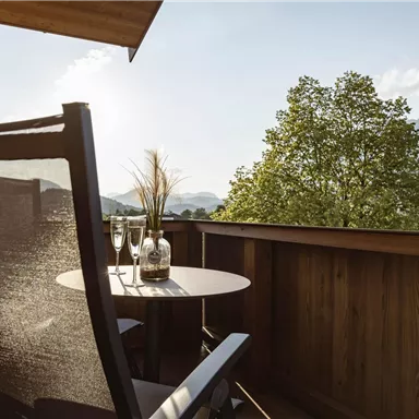 Ein gemütlicher Balkon mit einem Stuhl und einem kleinen Tisch. Im Hintergrund sind Bäume und Berge unter einem klaren Himmel zu sehen.