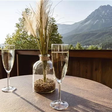Ein schöner Tisch mit zwei Gläsern und einer Vase auf einem Balkon. Im Hintergrund sind majestätische Berge und eine klare Himmel zu sehen.