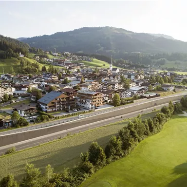 Eine malerische Landschaft mit einem kleinen Dorf im Tal und sanften Hügeln im Hintergrund. Die Straße und ein Golfplatz sind ebenfalls sichtbar.