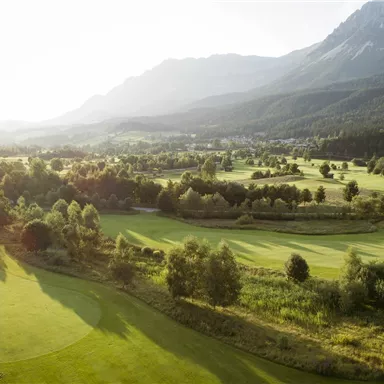 Eine weite Golfanlage umgeben von sanften Hügeln und Bäumen. Im Hintergrund sind Berge und ein klarer Himmel zu sehen.