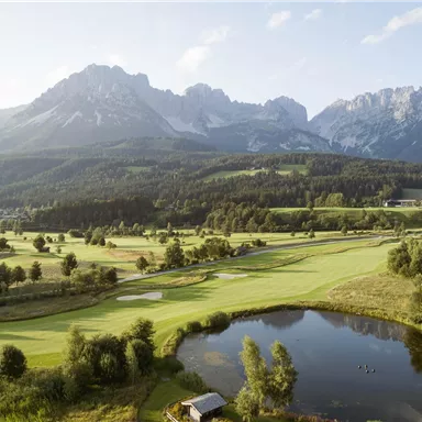 Eine malerische Golfanlage mit einem klaren Teich und sanften Hügeln. Im Hintergrund erheben sich majestätische Berge unter einem blauen Himmel.