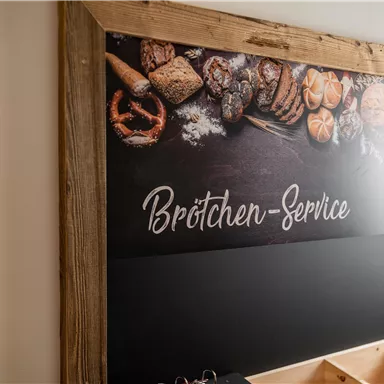 Ein Schild für den Brötchen-Service mit einer Auswahl an frischen Brötchen und Backwaren. Das Holzdesign verleiht dem Bild eine warme, einladende Atmosphäre.