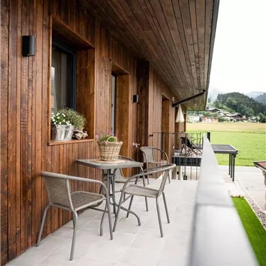 Eine gemütliche Terrasse mit Holzverkleidung und modernen Gartenmöbeln. Im Hintergrund sind Grünflächen und Berge sichtbar.
