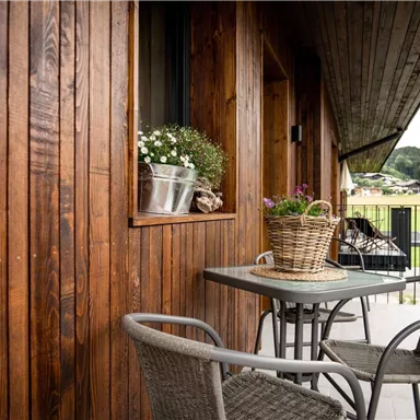 Ein gemütlicher Balkon mit Holzverkleidung und einem kleinen Tisch. Auf dem Tisch stehen Blumen in einem Korb und Töpfen.