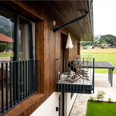 Eine moderne Holzfassade mit einem Balkon und gemütlichen Stühlen. Im Hintergrund sind grüne Wiesen und Berge zu sehen.