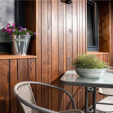 Ein gemütlicher Balkon mit einem kleinen Tisch und zwei Stühlen. Auf der Fensterbank stehen Pflanzen und die Wand ist aus warmem Holz.