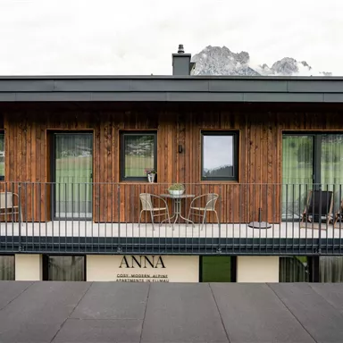Ein modernes Holzhaus mit einem Balkon und einem Tisch sowie Stühlen. Im Hintergrund sind Berge zu sehen.