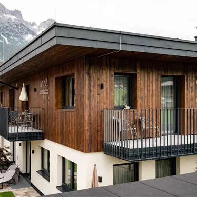 Ein modernes Holzhaus mit Balkonen und klaren Fenstern in den Bergen. Im Hintergrund sind majestätische Berge zu sehen.