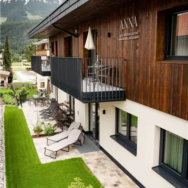 Ein modernes Gebäude mit Holzfassade und Balkon. Im Hintergrund sind grüne Wiesen und Berge zu sehen.