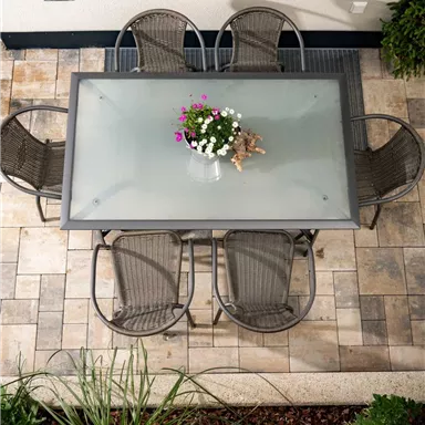 Ein schöner Tisch mit sechs Stühlen auf einer Pflasterfläche. Auf dem Tisch steht ein Topf mit bunten Blumen.