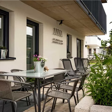 Eine moderne Terrasse mit einem Tisch und Stühlen. Im Hintergrund sind Pflanzen und eine elegante Fassade zu sehen.