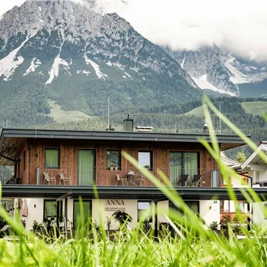 Ein modernes Haus mit Holzfassade, umgeben von grünem Gras. Im Hintergrund erheben sich majestätische Berge unter einem wolkigen Himmel.