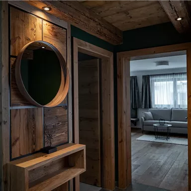 Ein gemütlicher Flur mit Holzwänden und einem runden Spiegel. Im Hintergrund ist ein heller Wohnraum mit einem Sofa und großen Fenstern zu sehen.