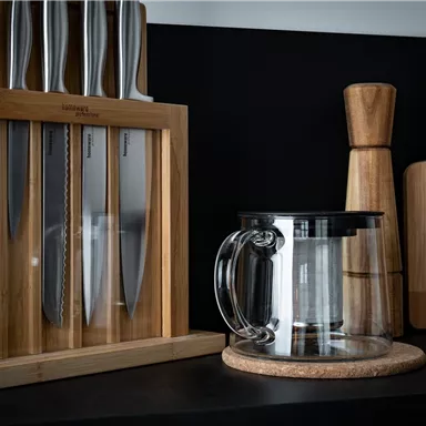 Ein Küchenbereich mit einem modernen Messerset in einem Holzblock. Darunter steht eine transparente Teekanne und einige Holzbrettchen.