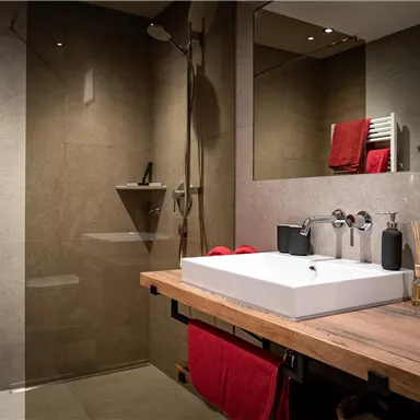 Ein modernes Badezimmer mit einer großen Dusche und einem eleganten Waschbecken. Rote Handtücher sorgen für einen Farbtupfer im stilvollen Design.