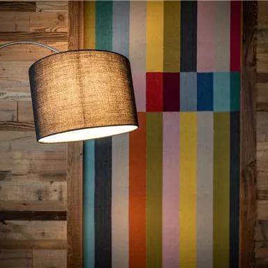 Eine Stehlampe steht vor einer bunten Wand mit streifenartigen Mustern. Der Hintergrund besteht aus Holzpaneelen, was eine gemütliche Atmosphäre schafft.