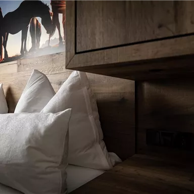 Ein gemütliches Schlafzimmer mit vielen weißen Kissen und rustikalen Holzmöbeln. Im Hintergrund ist ein Bild von Pferden, das dem Raum eine entspannte Atmosphäre verleiht.