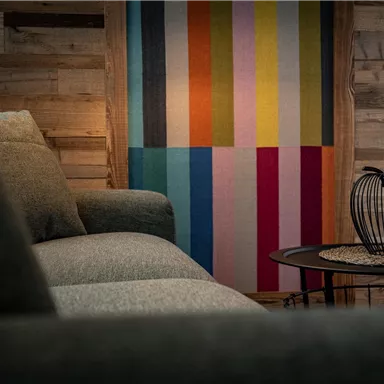 Ein gemütliches Wohnzimmer mit einem grauen Sofa und einer bunten Wanddekoration. Auf einem kleinen Tisch steht eine dekorative Drahtvase.