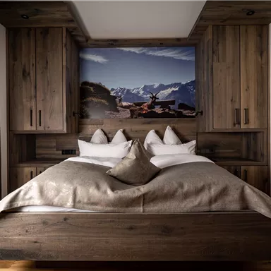 Ein modernes Schlafzimmer mit einem holzfarbenen Bett und eleganter Bettwäsche. Im Hintergrund befindet sich ein Bild von einer Berglandschaft.