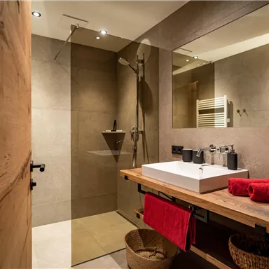 Ein modernes Badezimmer mit einer klaren Dusche und einer Holzablage. Es gibt ein Waschbecken und einige dekorative Elemente wie Handtücher und einen Spiegel.