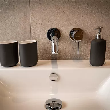 Ein modernes Waschbecken mit minimalistischen Accessoires. Zwei schwarze Becher und ein Seifenspender stehen neben dem Wasserhahn.