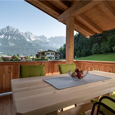 Eine gemütliche Terrasse mit einem Holzstuhl und einem Tisch, der mit einer Obstschale dekoriert ist. Im Hintergrund sind majestätische Berge und eine grüne Wiese zu sehen.