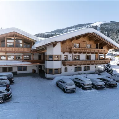 Ein traditionelles Gebäude in den Alpen, umgeben von schneebedeckten Tannen. Im Vordergrund stehen mehrere Autos auf dem verschneiten Parkplatz.