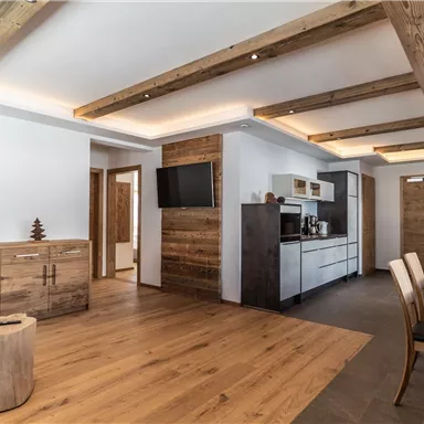 Ein modernes Wohnzimmer mit Holzbalken und Holzfußboden. Es gibt eine kleine Küche und eine Esstischgruppe.