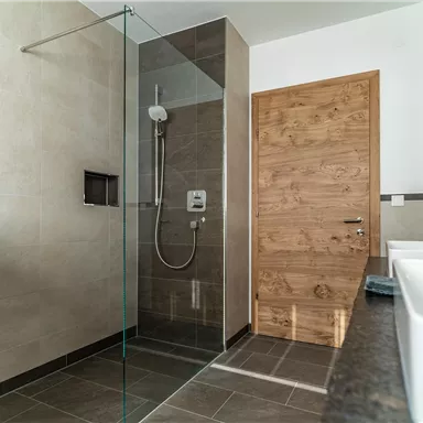Ein modernes Badezimmer mit einer gläsernen Duschkabine und elegantem Design. Die hellen Wände und der Holzton der Tür sorgen für eine angenehme Atmosphäre.