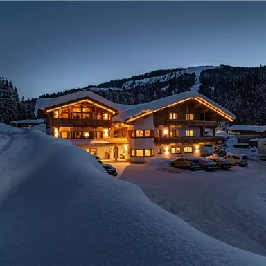 Ein gemütliches Chalet im Schnee, beleuchtet von warmem Licht. Die Umgebung ist von Bäumen und Bergen umgeben.