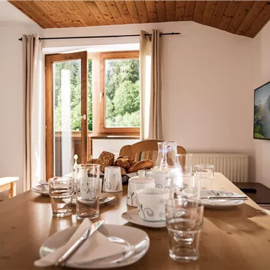 Ein gemütlicher Essbereich mit einem gedeckten Tisch und großen Fenstern, die viel Tageslicht hereinlassen. Im Hintergrund ist ein Fernseher sichtbar und die Einrichtung ist einladend und modern.