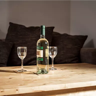 Eine Flasche Weißwein steht auf einem Holztisch, umgeben von zwei Weingläsern. Im Hintergrund sind ein paar dunkle Sofakissen zu sehen.