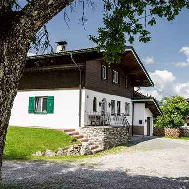 Ein schönes, modernes Haus mit grünen Fensterläden in einer ländlichen Umgebung. Es steht in der Nähe einer Straße, umgeben von Bäumen und Bergen.