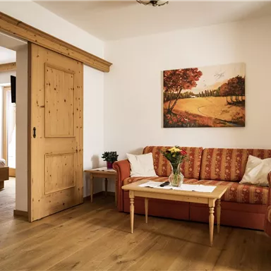 A cozy living space with a red sofa and a table. A landscape painting hangs on the wall, and there is a door leading to a bedroom.